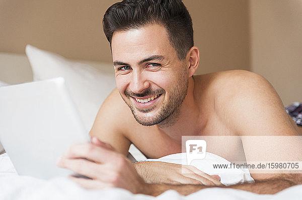 Glücklicher junger Mann  der ein digitales Tablet hält  während er zu Hause im Bett liegt