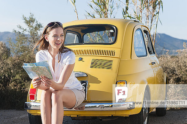 Porträt einer lachenden Frau mit Landkarte  die auf der Stoßstange eines gelben Weinautos sitzt  Sardinien  Italien