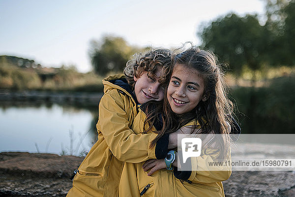 Junge umarmt lächelnde Schwester in gelben Regenmänteln am Flussufer