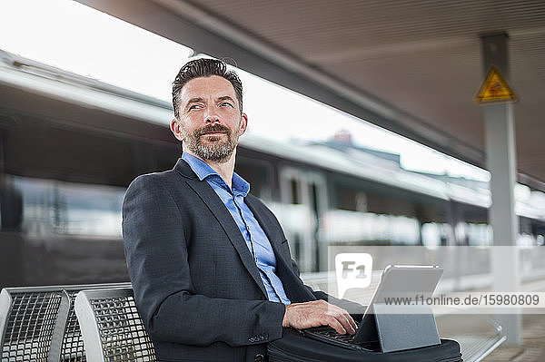 Confident businessman looking away while sitting and waiting with digital tablet at railroad station