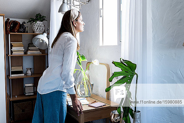 Woman looking through window while standing at desk in bedroom