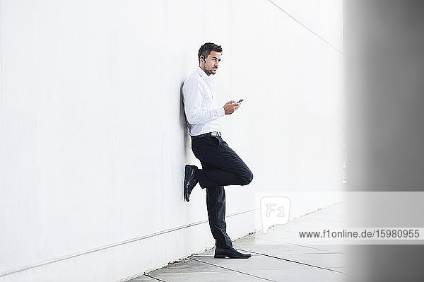 Geschäftsmann mit Smartphone an eine weiße Wand gelehnt
