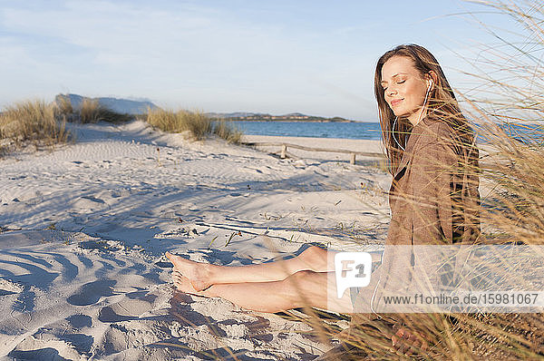 Portrait of woman sitting at beach dunes listening music with earphones  Sardinia  Italy