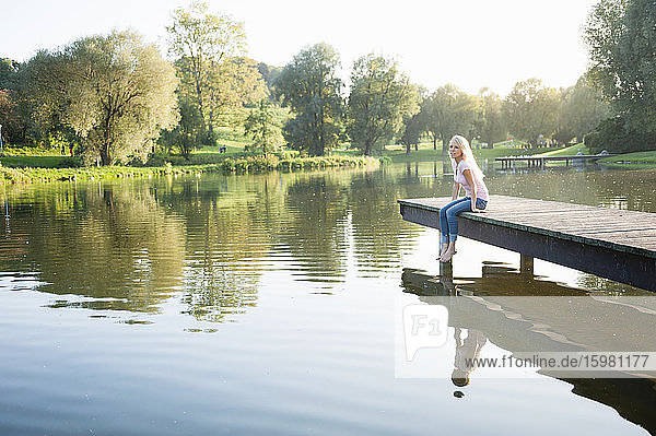 Junge Frau sitzt bei Sonnenuntergang auf einem Steg über einem See im Park