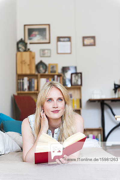Porträt einer lächelnden blonden Frau  die mit einem Buch auf der Couch liegt und in die Ferne schaut
