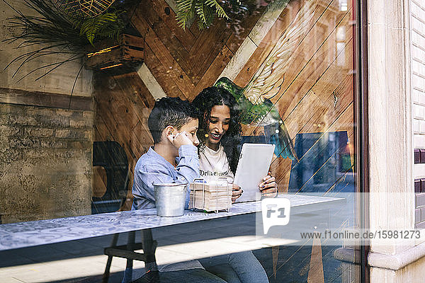 Lächelnde Frau  die mit ihrem Sohn im Restaurant sitzt und ein digitales Tablet benutzt  vom Fenster aus gesehen