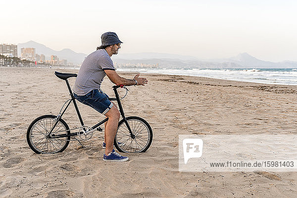 Älterer Mann mit Fahrrad  der am Strand sitzt und das Meer beobachtet