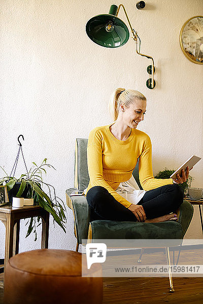 Lächelnde blonde Frau  die zu Hause auf einem Sessel sitzt und ein digitales Tablet benutzt
