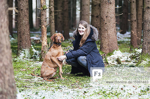 Lächelnde junge schöne Frau  die mit ihrem Hund vor einem Baum im Wald kauert