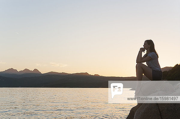 Frau sitzt bei Sonnenuntergang auf einem Felsen und blickt auf das Meer  Sardinien  Italien
