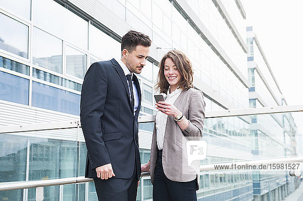Glückliches Geschäftspaar benutzt ein Smartphone  während es vor einem Bürogebäude steht