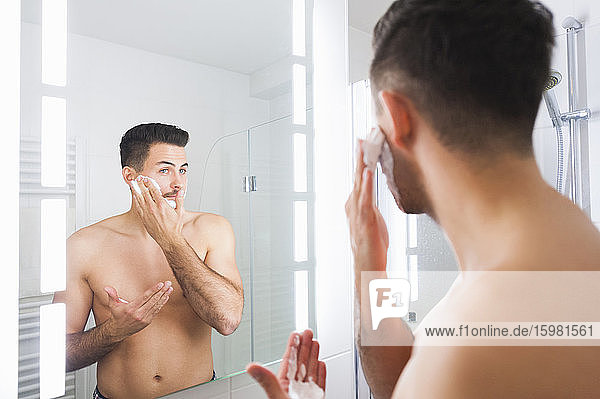 Spiegelung eines jungen Mannes  der zu Hause Rasierschaum auf die Wange aufträgt
