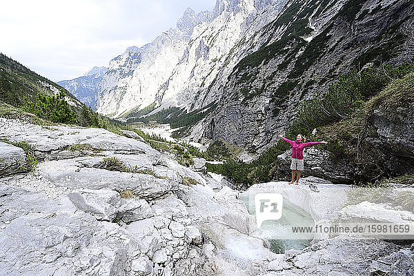 Weibliche Touristin steht mit ausgestreckten Armen auf einem Felsen am Bach vor den Bergen