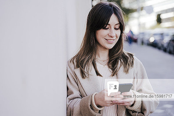 Porträt einer lächelnden jungen Frau  die an der Wand lehnt und auf ihr Smartphone schaut
