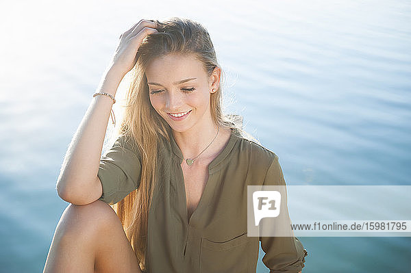 Porträt einer lächelnden jungen Frau  die bei Sonnenaufgang vor einem See sitzt