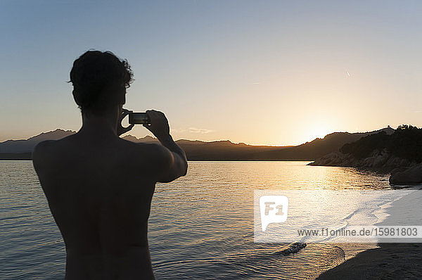 Rückansicht eines Mannes  der mit seinem Handy den Sonnenuntergang fotografiert  Sardinien  Italien