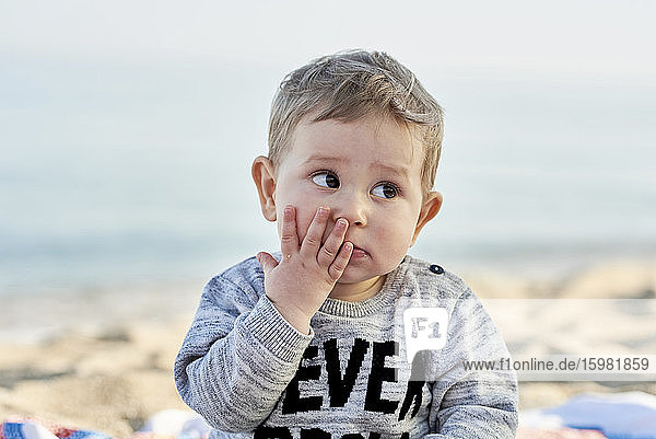 Close-up of cute boy with hand on face sitting at beach against sky