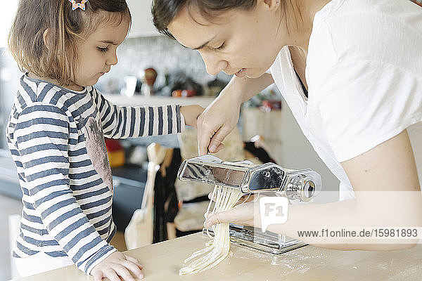 Cute girl looking at mother making spaghetti from equipment in kitchen