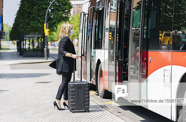 Geschäftsfrau mit Schutzmaske vor einem Bus mit Trolley-Tasche  Spanien
