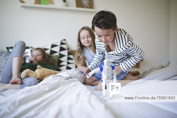 Excited little boy crouching on bed playing with toy robot while his sisters relaxing in the background