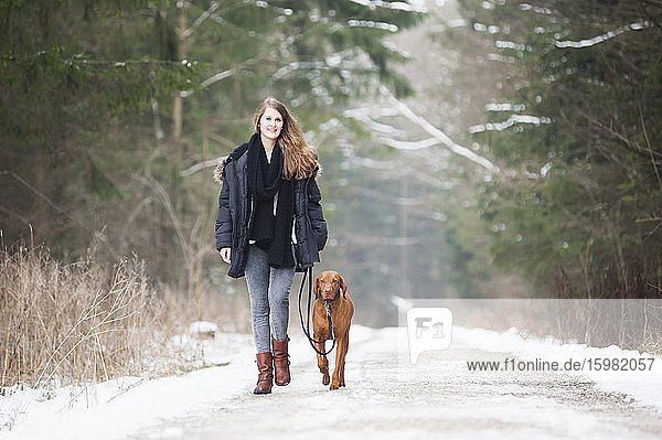 Schöne junge Frau geht mit Hund auf der Straße inmitten von Bäumen im Wald im Winter