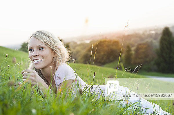 Smiling thoughtful young woman lying on grassy land against clear sky in park during sunset