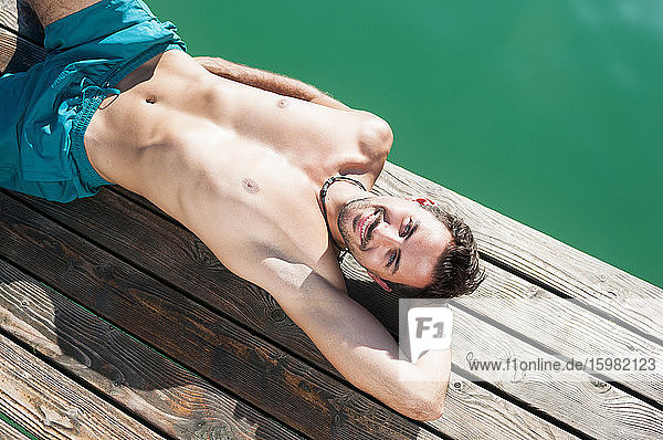 Porträt eines lächelnden jungen Mannes in Badeshorts beim Sonnenbaden auf einem Steg