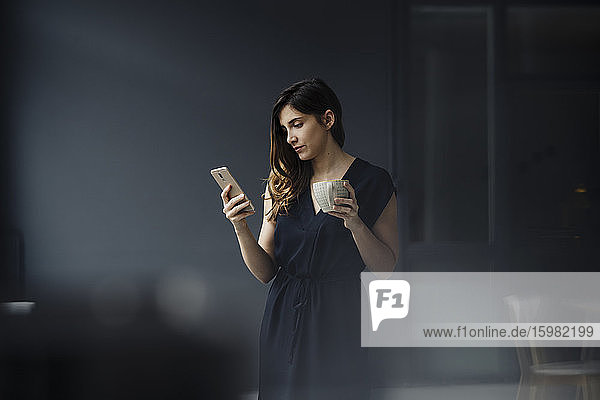 Portrait of serious young woman with cup of tea in a loft looking at cell phone