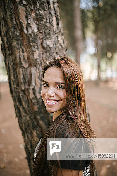 Seitenansicht Porträt einer lächelnden jungen Frau  die an einem Baumstamm im Wald steht