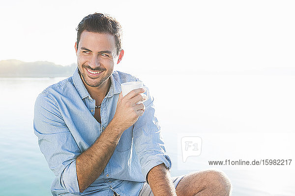 Porträt eines jungen Mannes  der sich mit einer Tasse Kaffee vor einem See entspannt