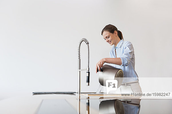 Lächelnde Frau im mittleren Erwachsenenalter  die Wasser in ein modernes Spülbecken gießt  während sie an der Wand steht