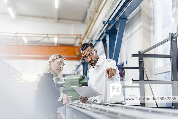 Geschäftsmann und Frau mit Papieren untersuchen Metallstangen in einer Fabrikhalle