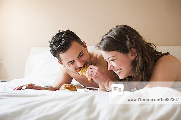 Glückliche junge Frau füttert einen Mann mit einem Croissant  während sie zu Hause im Bett liegt