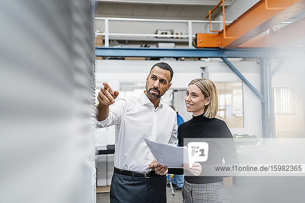 Geschäftsmann und Frau mit Papieren an einer Maschine in einer Fabrikhalle