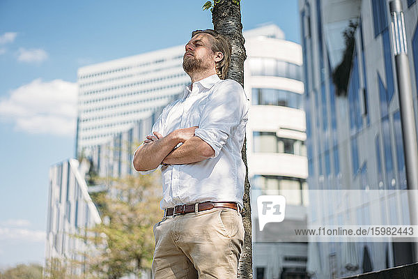 Businessman having a break in the city leaning against a tree trunk