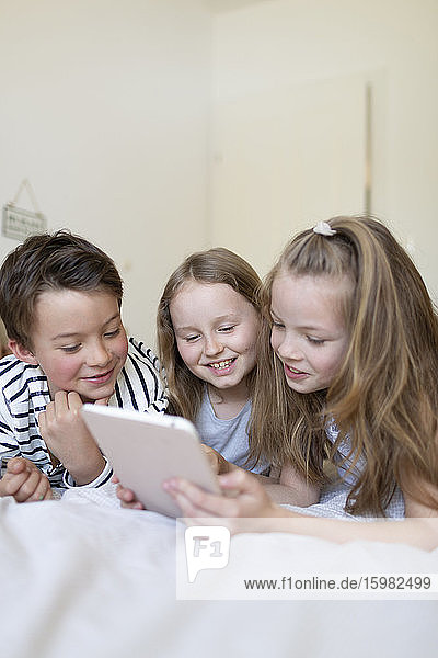 Portrait of boy and his two sisters lying on bed using digital tablet for homeschooling