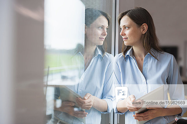 Nachdenkliche erwachsene Frau  die ein digitales Tablet hält und durch ein Fenster zu Hause schaut