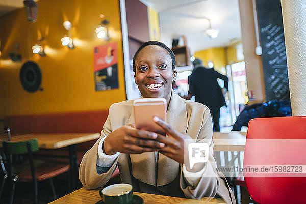 Happy young woman holding smart phone while sitting in coffee shop