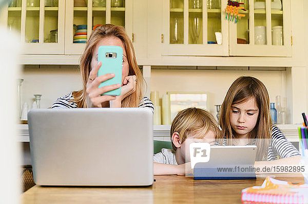 Frau hält ein Smartphone vor den Laptop  während Junge (4-5) und Mädchen (6-7) ein digitales Tablet benutzen