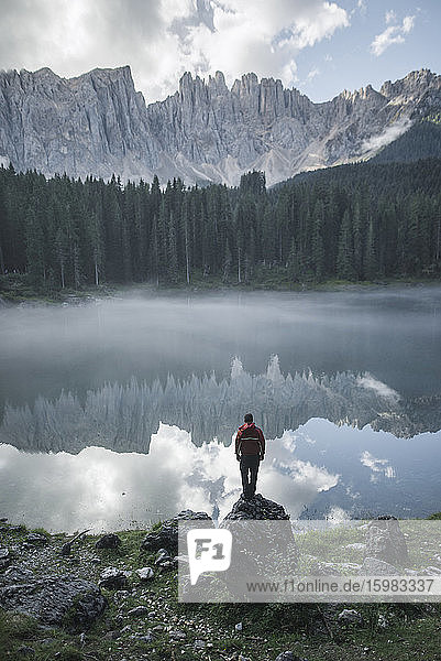 Italien  Carezza  Junger Mann steht in der Morgendämmerung auf Fels am Lago di Carezza in den Dolomiten