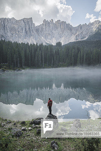 Italy  Carezza  Young man standing on rock at Lago di Carezza in Dolomite Alps at dawn