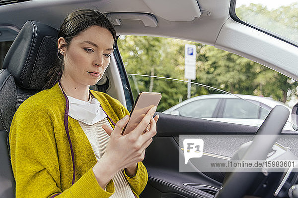 Portrait of serious woman with protective mask in car looking at smartphone