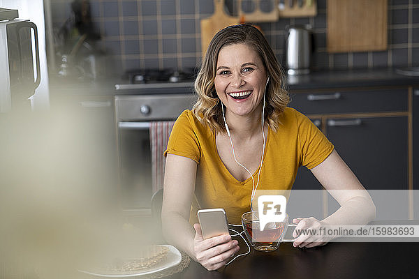 Porträt einer glücklichen Frau mit Smartphone und Kopfhörern  die zu Hause am Küchentisch sitzt