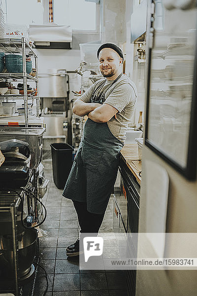 Smiling confident male owner standing with arms crossed in kitchen at restaurant