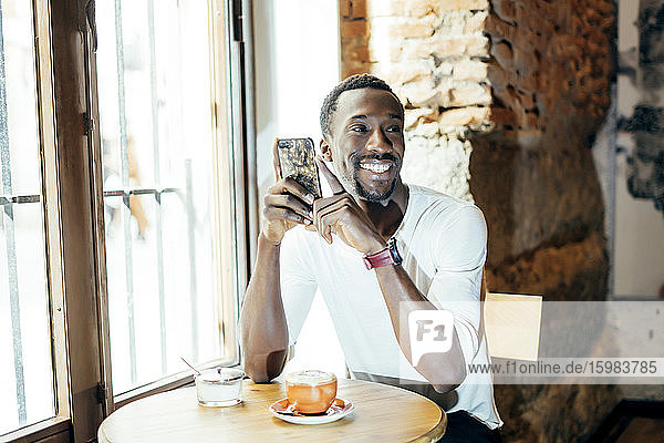 Smiling young man with coffee on table holding smart phone looking away while sitting in cafe