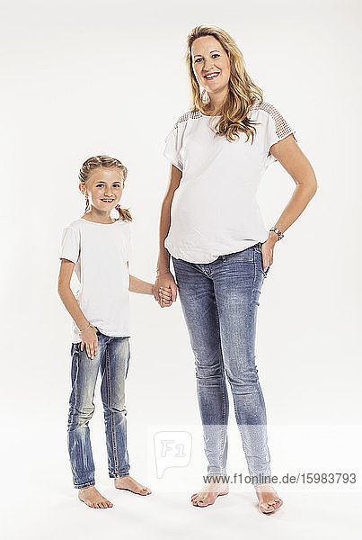 Portrait of happy pregnant woman standing hand in hand with her daughter in front of white background