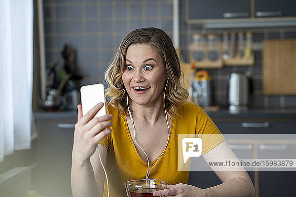 Surprised woman having a video chat with smartphone in kitchen at home