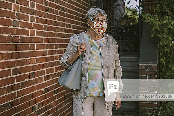 Retired senior woman wearing face mask while walking by brick wall during coronavirus epidemic