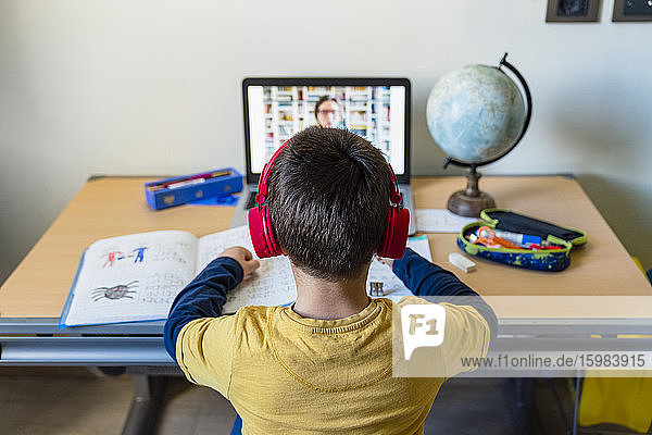 Rückansicht eines Jungen  der zu Hause während des Ausbruchs einer Pandemie per Videoanruf einem Lehrer zuhört