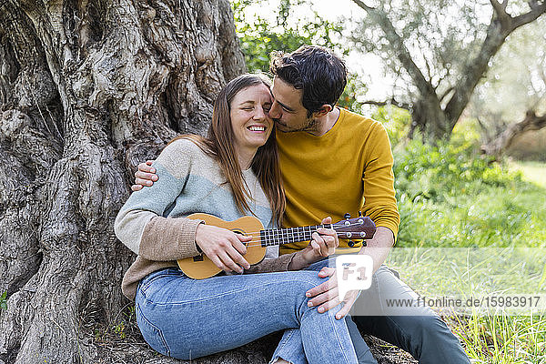 Romantischer Freund küsst glückliche Frau  die Gitarre spielt  während sie gegen einen Baumstamm sitzt